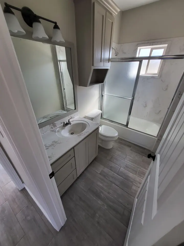 Completed Better Builders bathroom with a tub and shower enclosure, marble-look wall surround, gray vanity, mirror, and wood-look tile floor.