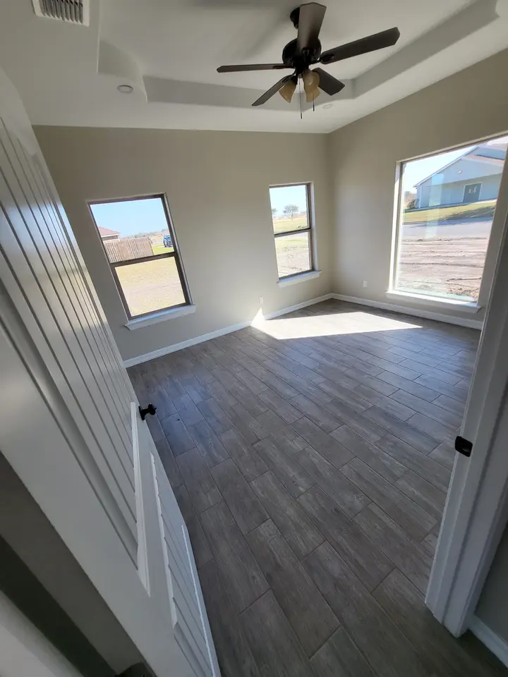 Completed Better Builders bedroom with a tray ceiling, ceiling fan, three tall windows, neutral walls, and wood-look tile flooring.