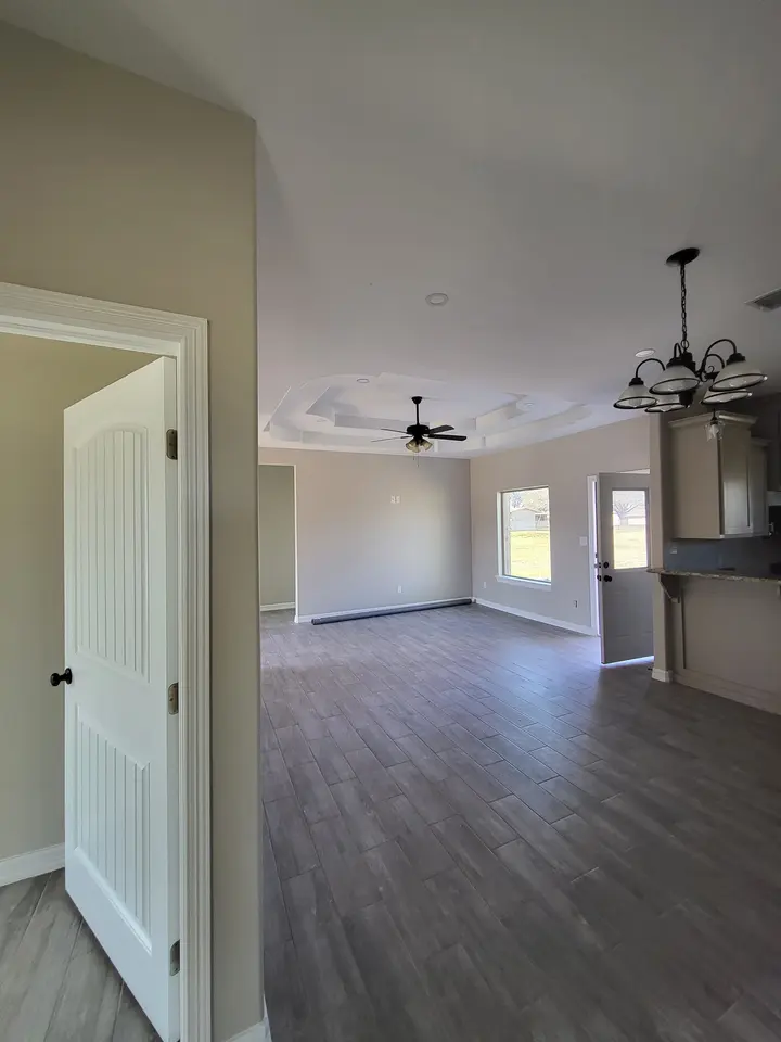 Completed Better Builders open living area with a decorative tray ceiling, ceiling fan, wood-look tile, tall windows, and a connection to the kitchen.