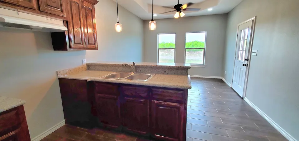 Finished Better Builders kitchen with dark wood cabinetry, granite counters, a double-basin sink, pendant lights, and an open living area.