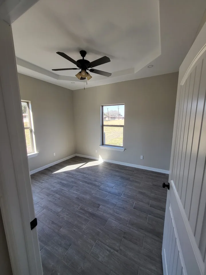 Finished Better Builders bedroom with a tray ceiling, black ceiling fan, two tall windows, neutral walls, and wood-look tile flooring.