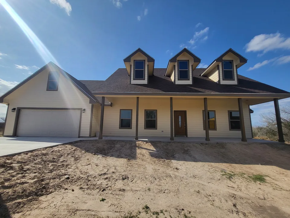 Front exterior of a completed Better Builders farmhouse-style home with three dormers, a full-width covered porch, neutral siding, and an attached garage.