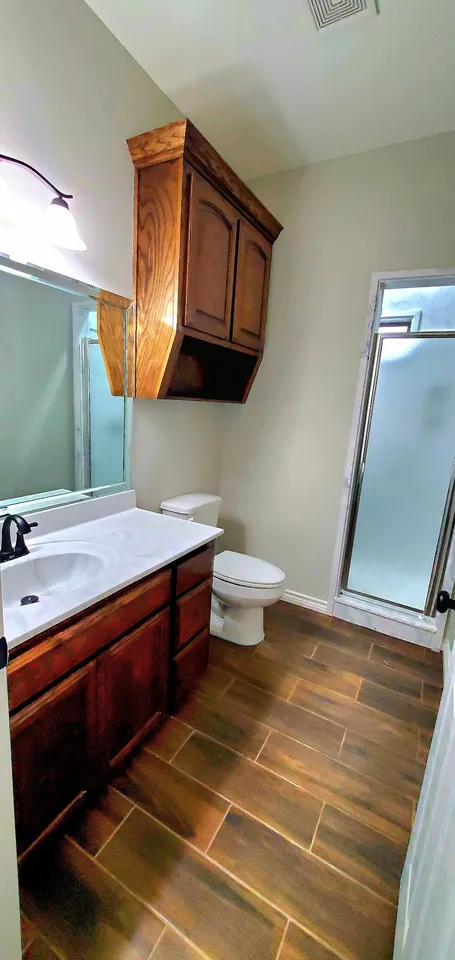 Finished Better Builders bathroom with a custom wood vanity, white counter, dark faucet, wall cabinet, toilet, wood-look tile, and frosted-glass shower enclosure.