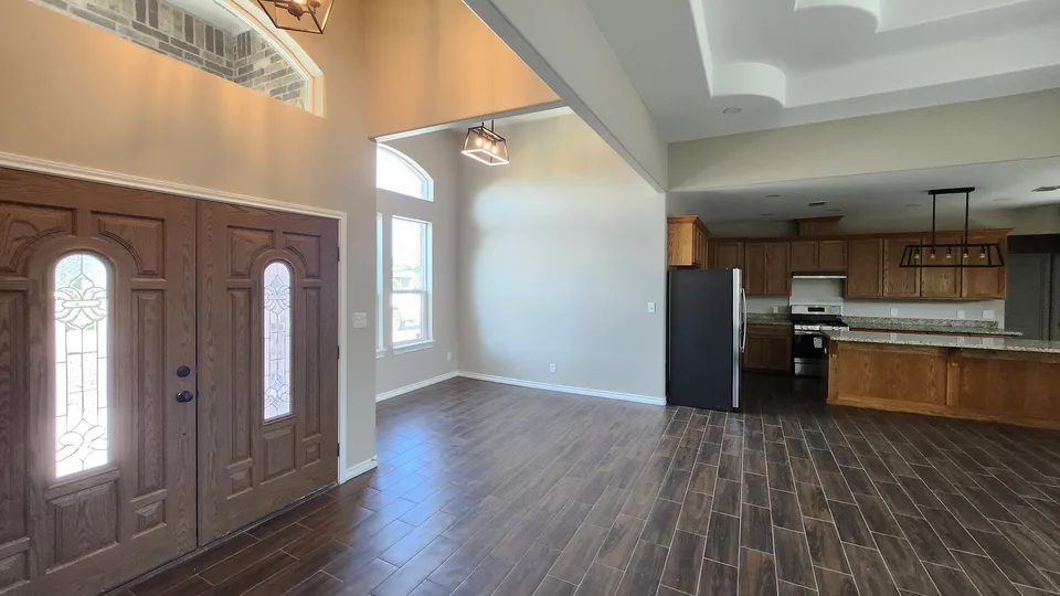 Finished Better Builders open-plan interior with decorative wood entry doors, tall windows, wood-look tile, a tray ceiling, and a wood-cabinet kitchen with granite counters.