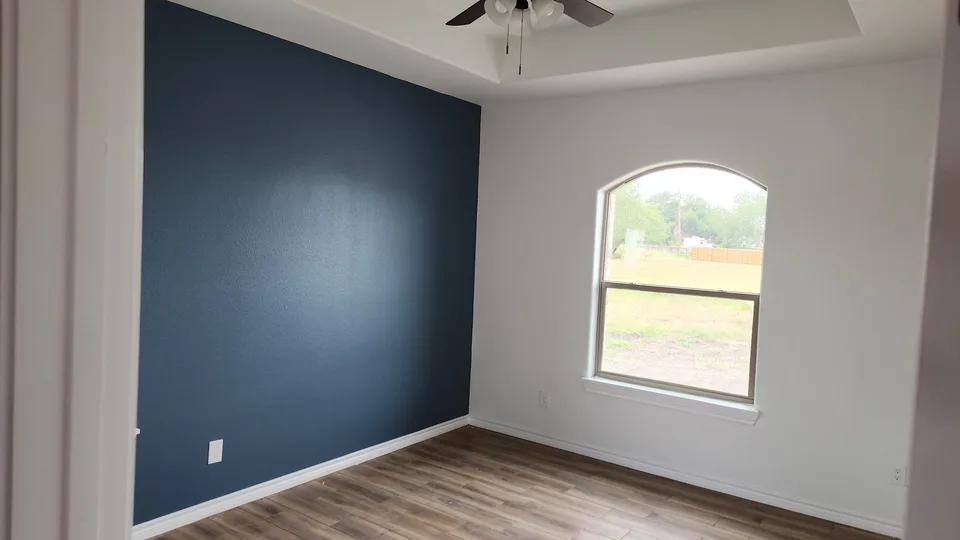 Finished Better Builders bedroom with a navy accent wall, tray ceiling, ceiling fan, arched window, white trim, and wood-look tile flooring.