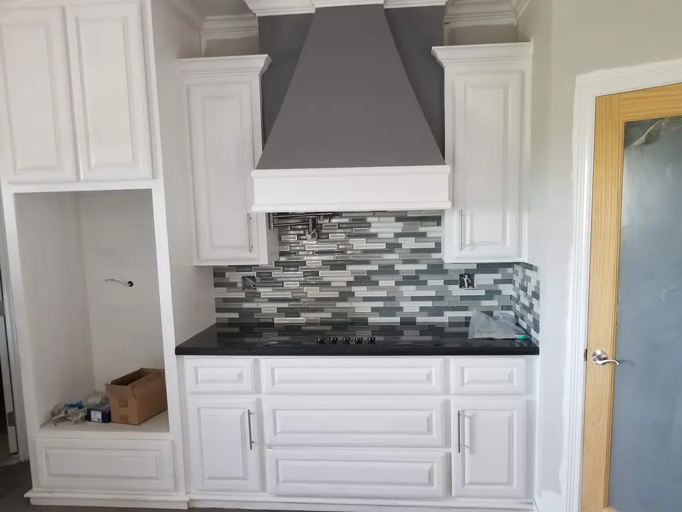 White custom kitchen cabinetry, decorative range hood, dark counter, and mosaic backsplash during finish work.