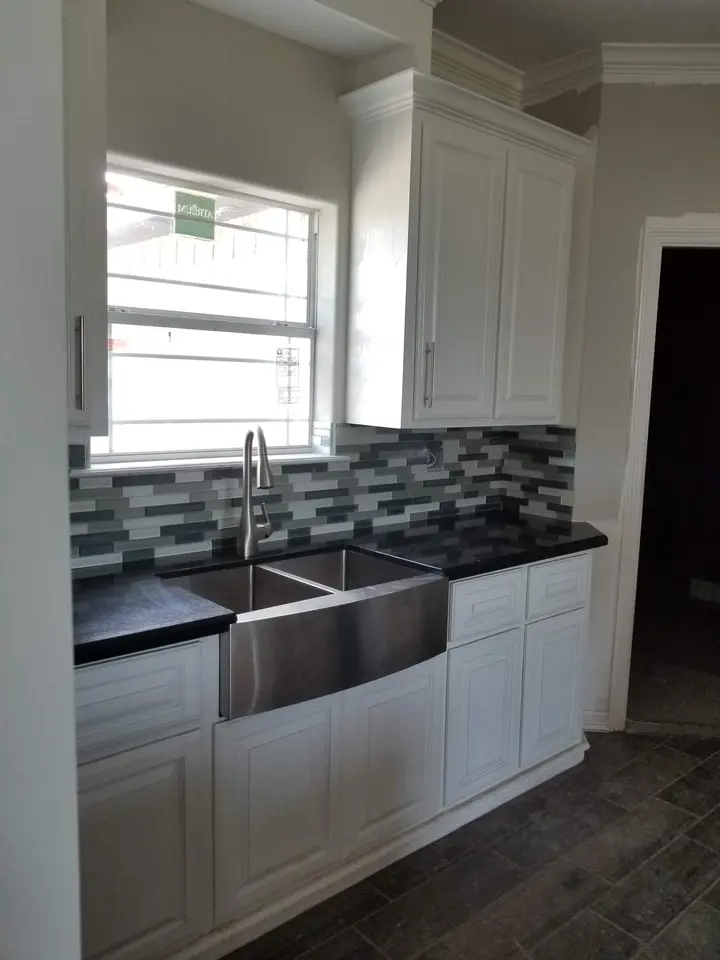 Stainless farmhouse sink set in white custom kitchen cabinetry with a mosaic backsplash and dark counter.