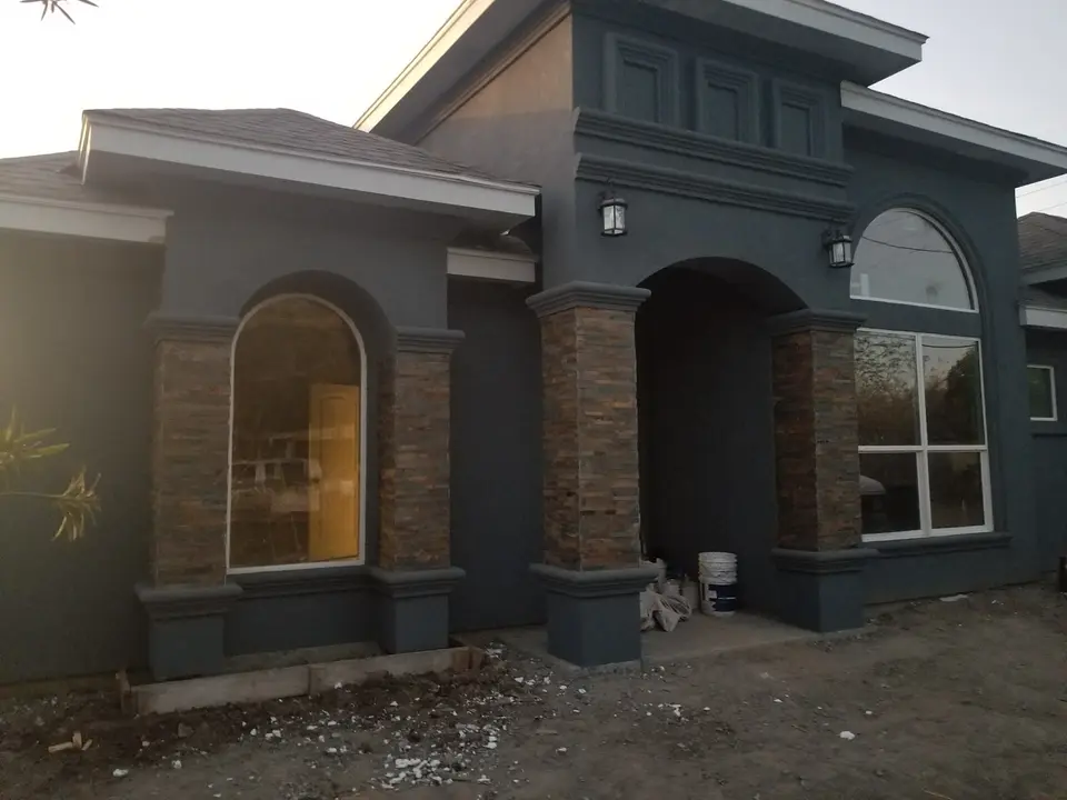 Dark stucco Better Builders home with stone entry columns, arched openings, and exterior finish work nearing completion.