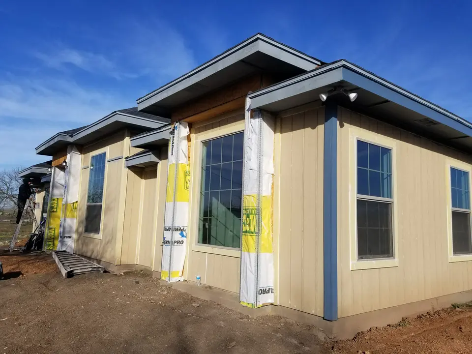Better Builders home with vertical tan siding, blue trim, deep roof overhangs, and installed windows during exterior finish work.
